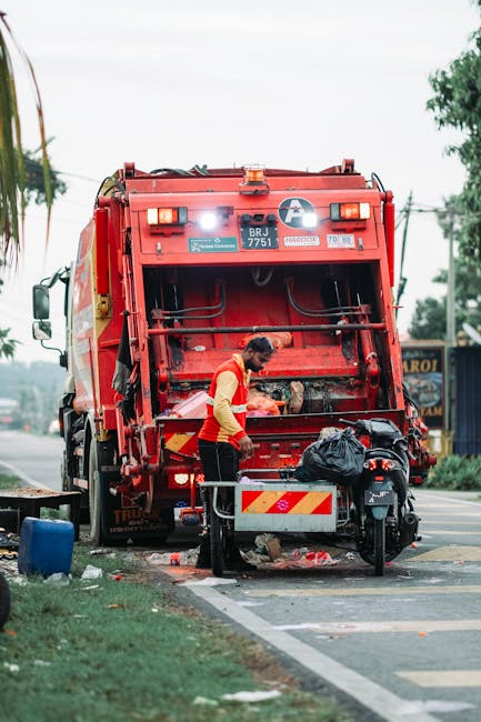 A sanitation worker wearing a high-visibility orange vest and dark trousers is operating a large red rubbish collection vehicle on an urban street. The rear section of the waste truck has an open hatc