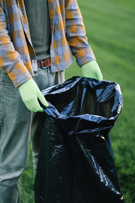 A sanitation worker wearing a high-visibility orange vest and dark trousers is operating a large red rubbish collection vehicle on an urban street. The rear section of the waste truck has an open hatc