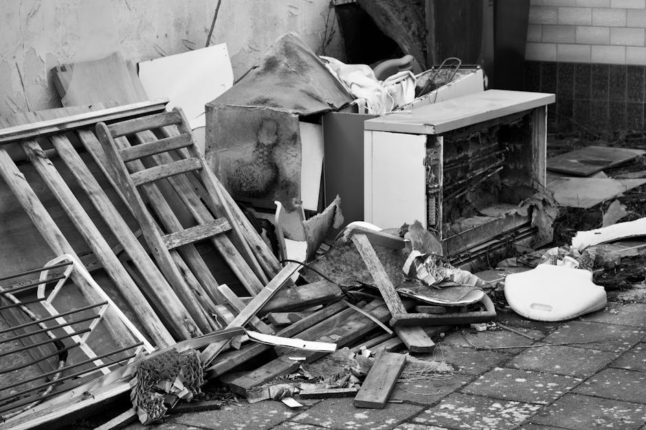 A pile of mixed construction debris and household waste is situated at the base of a multi-storey residential building, with rubble including bricks, broken concrete, and scraps of metal scattered acr
